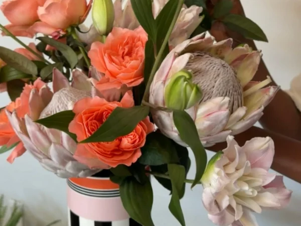 Floral Arrangement in Memphis vase Colorful bouquet of coral roses, peach blooms, pale protea, and lush green leaves in a tall striped vase; cheerful, elegant, and inviting.