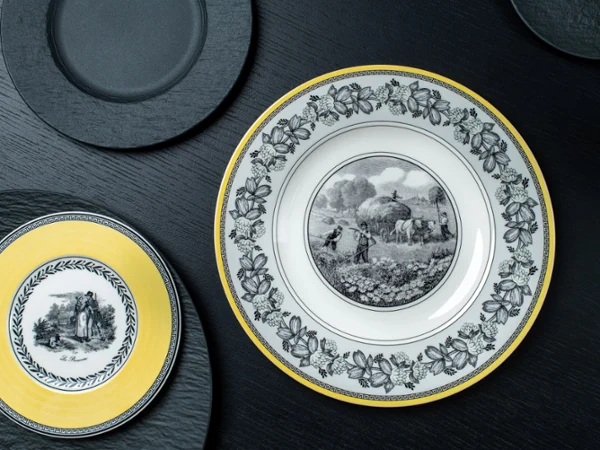 Audun and Manufacture Rock plates on a black table Elegant plates on a dark surface; one large with a rural scene and floral border, another smaller with a yellow rim depicting a couple.
