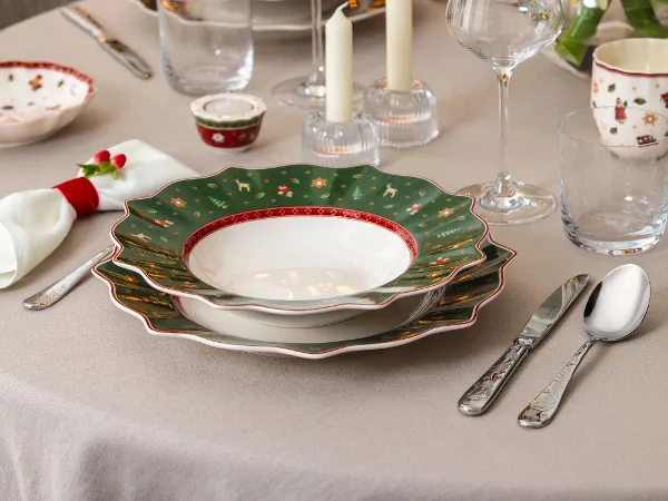 Toys Delight 2025 Collection, Christmas decorated. A festively set dining table with Christmas-decorated plates, glasses, candles, and flowers in front of a decorated Christmas tree.