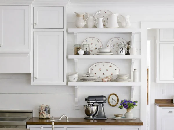 petite fleur items on a kitchen self as art White kitchen with open shelves displaying vintage floral plates, white pitchers, jars, and a framed picture. A mixer and flowers sit on the counter.