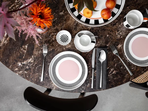 Colour explosion with Memphis by Villeroy & Boch A round marble table with two Memphis place settings, each with a plate, cutlery and a cup. A bowl of apples and a Memphis vase with flowers adorn the center.