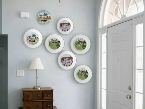 design naif plates on a wall as art Wall-mounted decorative plates with countryside scenes arranged above a wooden chest and lamp. Soft lighting from a nearby window creates a cozy entryway.