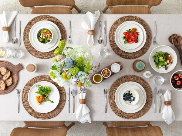 Vista dall'alto di una tavola da pranzo splendidamente apparecchiata con insalate, frutta, pane, bicchieri d'acqua e fiori al centro su una tovaglia beige.