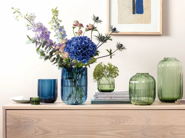 Fleur vases on a brown dresser Green and blue glass vases in various shapes some with flowers in them standing on wood furniture with a picture on the wall behind them