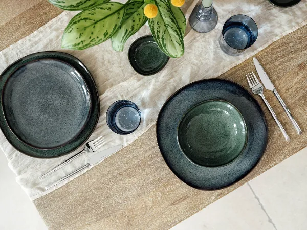 Lave place settings with green plates, cutlery and blue glasses decorate a wooden table Two Villeroy & Boch Lave place settings with green plates, cutlery and blue glasses adorn a wooden table with a white table runner. In the centre is a green-leaved plant in a vase.