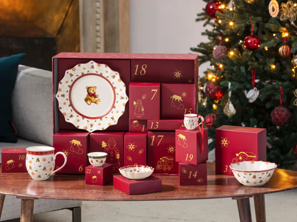 One red advent calendar and some Toys Delight ceramic articels like a mug plate or a bowl standing on a wooden table next to a decorated Christmas tree.