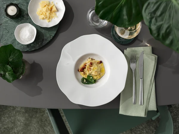 A white Pura pasta plate with pasta and side dishes on a grey table A white Pura pasta plate with pasta and side dishes on a grey table, next to it cutlery, a green napkin and a small bowl of cheese.