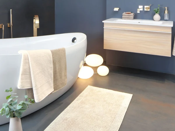 Biała wanna Villeroy & Boch z drewnianymi meblami łazienkowymi Nowoczesna łazienka z białą wanną wolnostojącą, umywalką wiszącą, złotymi bateriami, ręcznikami, rośliną doniczkową i podświetlanymi kulami na podłodze