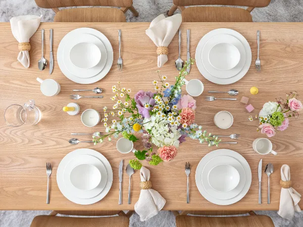 For Me in Spring A wooden dining table set for four people with white plates, cups, and cutlery. In the centre, there is a floral arrangement with pink and white flowers. Glass jugs are ready, and rolled napkins are placed next to the plates.