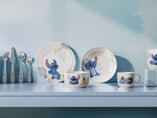 Una serie de platos y tazas de la colección Disney Stitch con motivos de dibujos animados, expuestos en una estantería azul claro junto con otros utensilios.