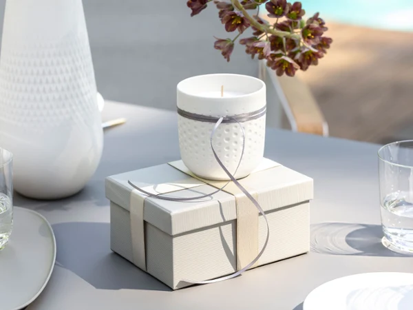 Una vela Manufacture Blanc con un lazo gris está sobre una caja de regalo cerrada en una mesa Una vela blanca Manufacture Blanc con un lazo gris está sobre una caja de regalo cerrada en una mesa. Cerca hay dos vasos de agua y un jarrón Manufacture Blanc.