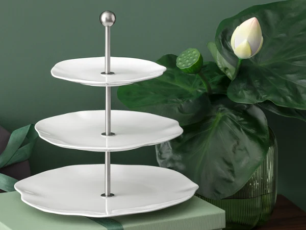 Three-tiered white Pura étagère with metal handle, next to it a large green lotus plant and a closed gift box.