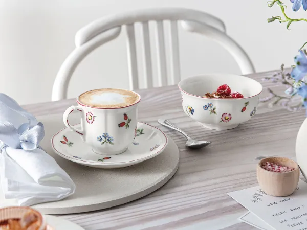A cup of cappuccino and a bowl of muesli on a table set with a napkin, a spoon, and a flower vase