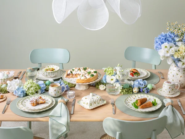 A pastel-themed dining table set for four, featuring plates of food, teacups, blue and white floral arrangements, and elegant tableware, creating a fresh and inviting atmosphere.
