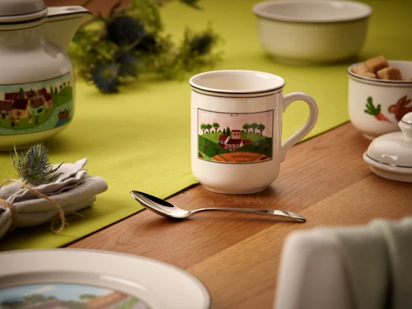A mug with a countryside illustration on a wooden table. Surrounded by a teapot, spoon, and napkin on a green tablecloth. Cozy and inviting setting.