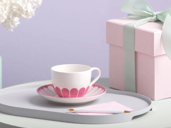A pastel-themed scene with a teacup and saucer featuring a pink pattern on a gray tray. Nearby is a pink gift box with a mint green ribbon, set on a lilac surface.