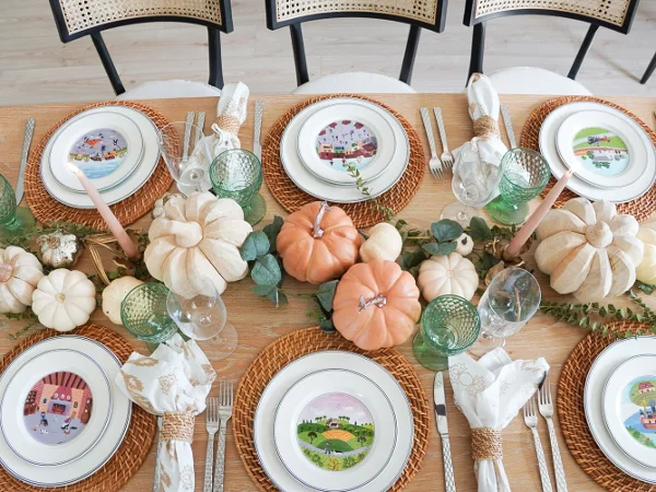 A wooden dining table set for a meal with decorative pumpkins, green glasses, white plates with illustrated scenes, and natural woven placemats.