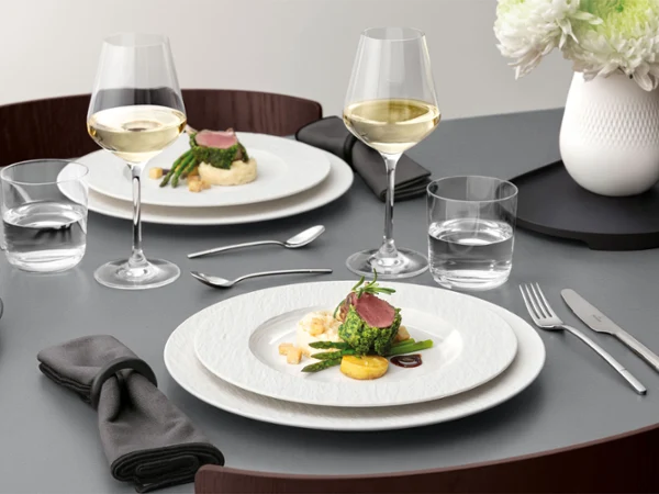 A formal table setting with a Manufacture plate on top of a plate A formal table setting with a white plate on a white plate with food on it, glassware, and silver cutlery on a grey table.
