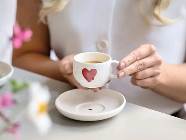 Taza de café y platillo de with love con motivo de corazones rojos Una persona sostiene una taza de café blanca con un motivo de corazones rojos sobre un platillo a juego con motivo de corazones rojos sobre una mesa.