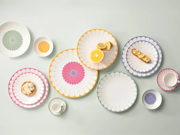Various colourful, patterned plates, bowls and a cup are arranged on a bright surface. Various colourful, patterned plates, bowls and a cup are arranged on a light-coloured surface, each with a different design. Pastries are placed on some of the plates.