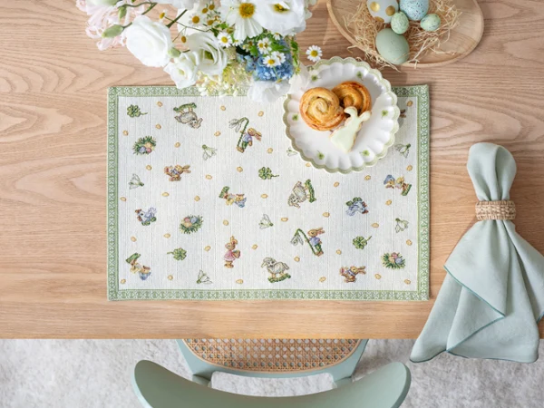 A wooden table set with a Easter Delight placemat A wooden table set with a Easter Delight placemat, pastries on a plate, flowers, dyed eggs, and a folded napkin.