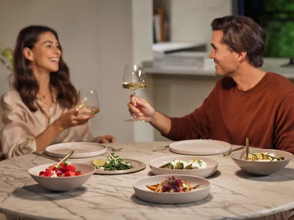 Un couple est assis à une table ronde dressée avec La Boule Beige de Villeroy & Boch, souriant et trinquant avec des verres à vin.