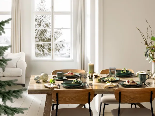 Lave Vert set for Christmas A wooden dining table for four with Lave Vert plates, cups, and candles in a bright room overlooking the snow outside.