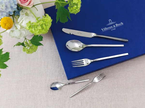 Set de couverts avec couteau, cuillère et fourchette sur un tissu bleu à côté d'une composition florale sur fond beige Set de couverts avec couteau, cuillère et fourchette sur un tissu bleu à côté d'une composition florale sur fond beige