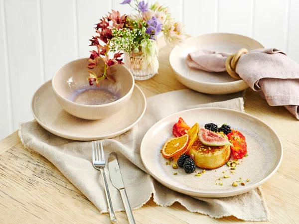 aus-perlemorsand-indoor-food-nopeople-dinnerset7069 The Perlemor Sand dinner set for one, featuring a plate filled with a dessert topped with fresh fruits, rests on a brown linen tablecloth atop a rustic wooden table, paired with silver cutlery.