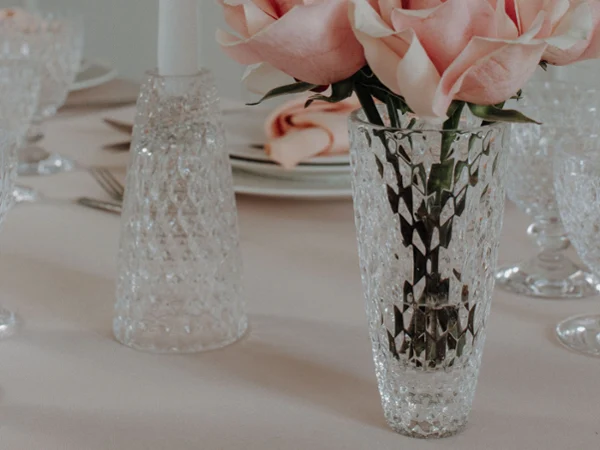 An elegant dining table setting with a bouquet of pink roses in a Boston vase next to a large candle in a Boston candleholder, together with Boston glasses