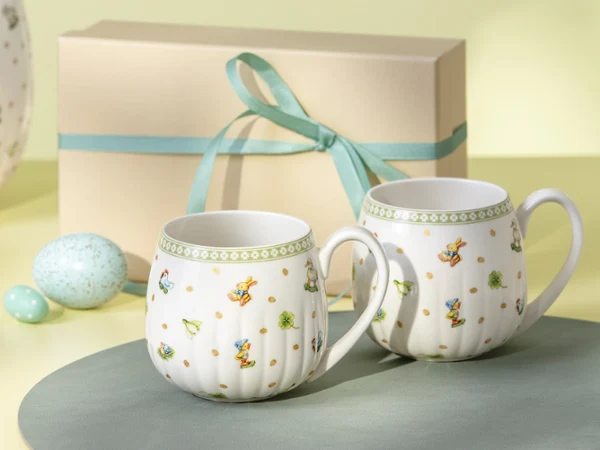 Zwei Ostern-Freude-Becher mit Blumenmustern stehen vor einer Geschenkbox, die mit einem hellblauen Band verziert ist.