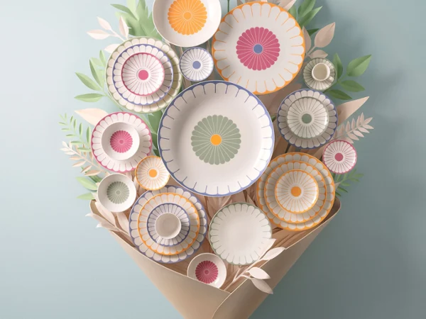 A floral arrangement of kitchenware with various plates and bowls decorated with fruit motifs, wrapped in brown paper and tied with a light blue ribbon, against a light blue background.