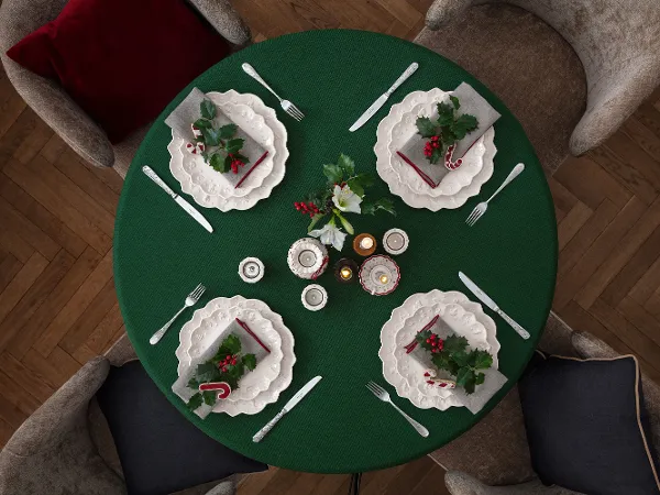 Un tavolo rotondo per quattro persone con piatti bianchi Toy's Delight Royal Classic di Villeroy & Boch, una tovaglia verde e decorazioni natalizie, tra cui foglie di agrifoglio, bacche e bastoncini di zucchero, visti dall'alto.