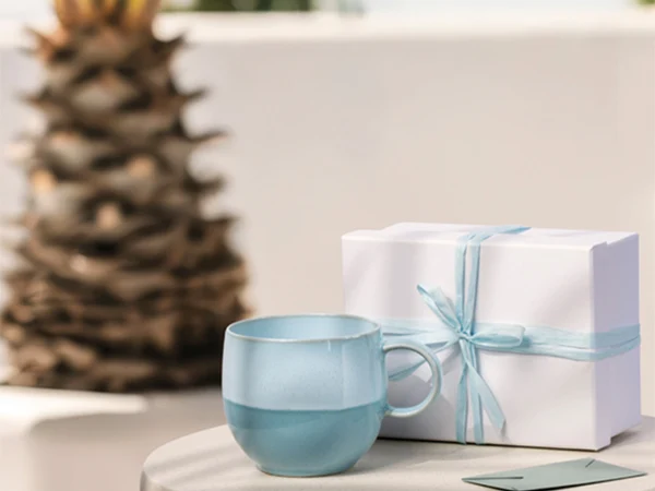 Taza Perlemor Aqua Una taza de cristal azul claro y una caja de regalo blanca con un lazo azul sobre una mesa redonda de piedra; al fondo, una palmera.