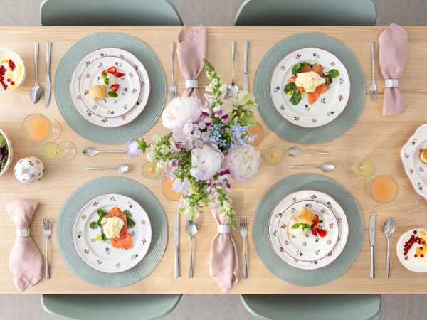 Ein gedeckter Esstisch für vier Personen mit Petite Fleur Blumentellern, rosa Servietten, Getränken und einem Blumenschmuck.