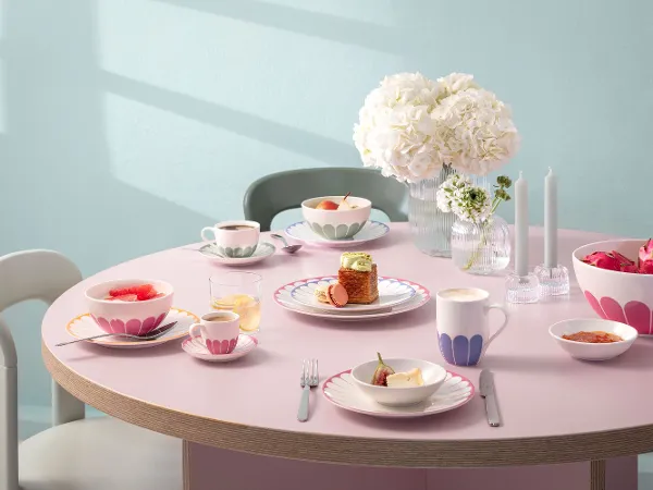 A round table with plates, cups and bowls full of food, decorated with pink patterns. A round table with plates, cups and bowls full of food, decorated with pink patterns. In the centre is a vase of white flowers and two unlit candles. Chairs surround the table.