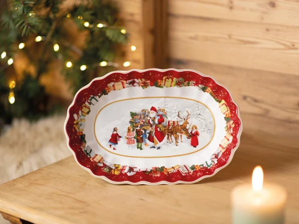 Toy's Fantasy Oval Platter on wood table with candle Festive ceramic plate with Santa, reindeer, and children in holiday outfits. Red border with gifts. Warm candlelight and fir branches in background. Cozy and joyful atmosphere.