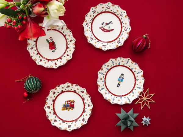 Quatre assiettes de Noël décoratives avec des motifs festifs sur une nappe rouge, entourées d'ornements et de fleurs.