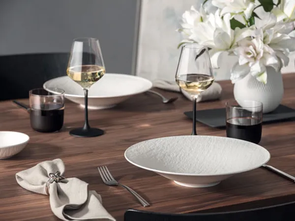 Manufacture dining table with plates, glasses and vase A dining table set with Manufacture white plates, wine glasses, cutlery, and a napkin. A Manufacture vase of white flowers is in the background.
