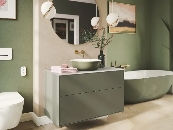 Kreatives grünes Bad mit Artis Waschbecken von Villeroy & Boch Modernes Badezimmer mit grünem Waschtisch und Badewanne, rundem Spiegel, Wandarmatur, weißem Waschbecken, Zierpflanzen und zwei Hängelampen. Im Hintergrund sind eine Badewanne und Wandkunst zu sehen.