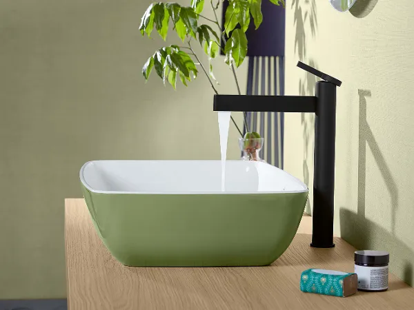 Svart Dawn-servantarmatur med grønn Avento-servant En moderne servant med grønn utside og hvit innside står på en treplate og har en svart kran med rennende vann. Ved siden av servanten står det et glass og en eske. En grønn plante i bakgrunnen.