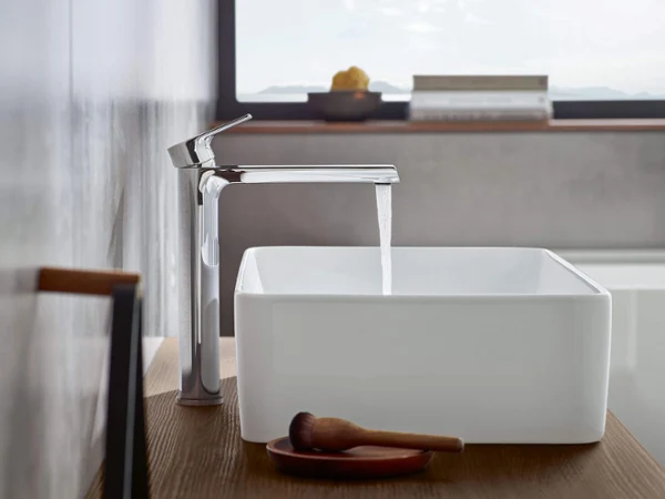 Liberty washbasin tap with a Collaro washbasin A modern chrome tap from which water is flowing into a rectangular white washbasin on a wooden shelf in a minimalist bathroom with a window in the background.