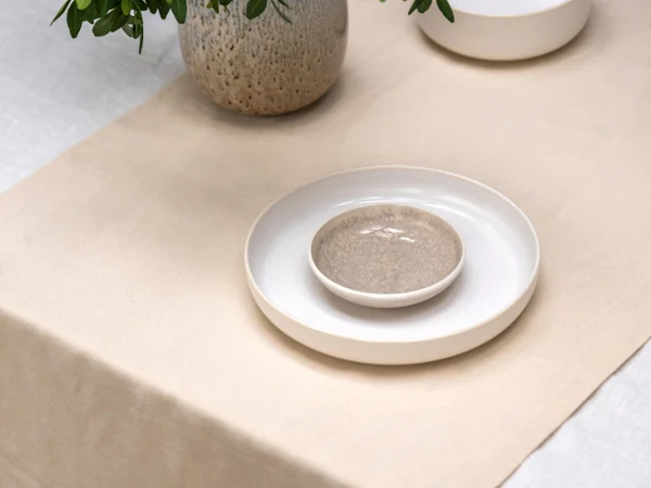 A table with a beige runner, a stack of ceramic plates, a bowl, and a vase with green leafy branches.

