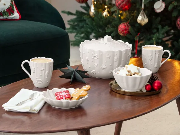 A decorated Christmas tree with red ornaments stands behind a coffee table set with mugs, cookies, and festive decorations in a cozy living room.