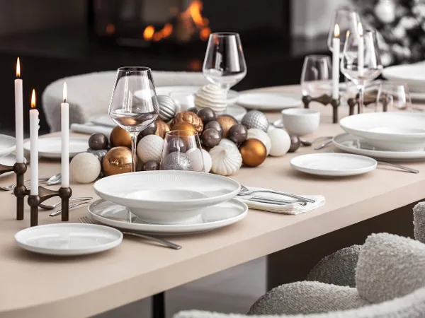 Elegant Christmas with Afina A dining table set with white Afina plates, glasses, candles, and golden Christmas decorations, with a fireplace in the background.
