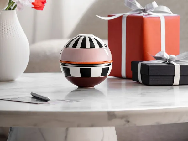 La petite Boule Memphis und zwei verpackte Geschenke stehen auf einem Tisch. La petite Boule Memphis von Villeroy & Boch und zwei verpackte Geschenke stehen auf einem Marmortisch, im Hintergrund steht eine Vase mit Blumen.