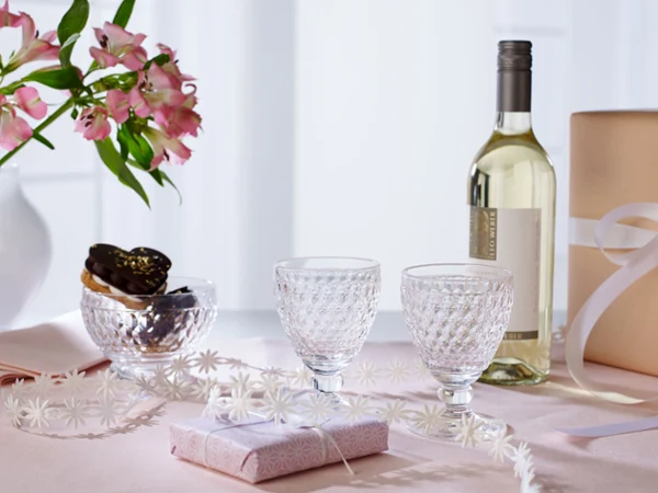 A table set with glasses, white wine, chocolates, a gift box, flowers and a paper garland.