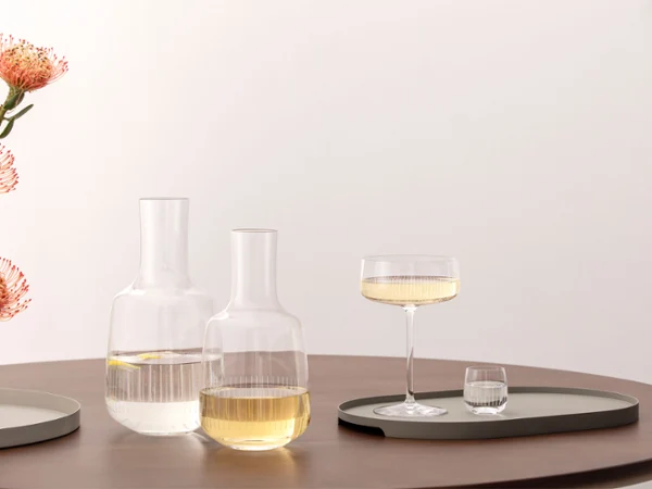 Two glass carafes, a champagne glass and a water glass from the Afina collection are placed on trays on a wooden table with orange flowers in a vase.
