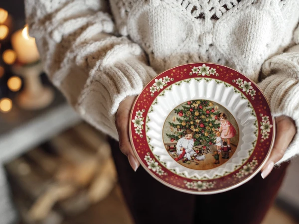 A person holding a Toys Fantasy Bowl A person wearing a cozy sweater holds a decorative plate with a Christmas scene of children near a tree. Warm, festive ambiance enhances holiday spirit.