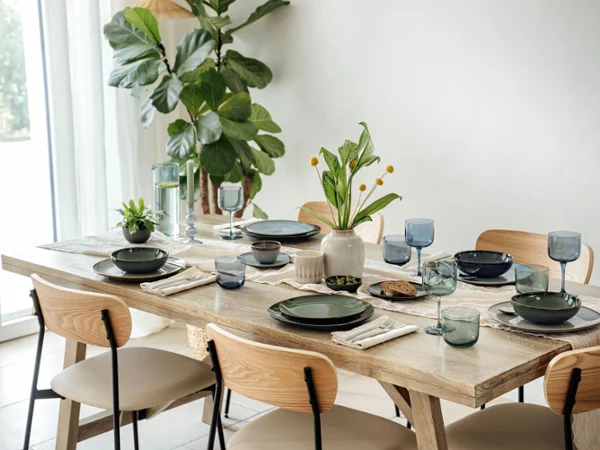 The dining table set with varios items from the Lave collection by Villeroy & Boch in green and grey, accentuated by blue glasses and napkins. A vase of yellow flowers and a small potted plant provide decoration, while a large plant stands by the window.